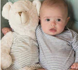 Baby with teddy bear wearing grey striped overalls.